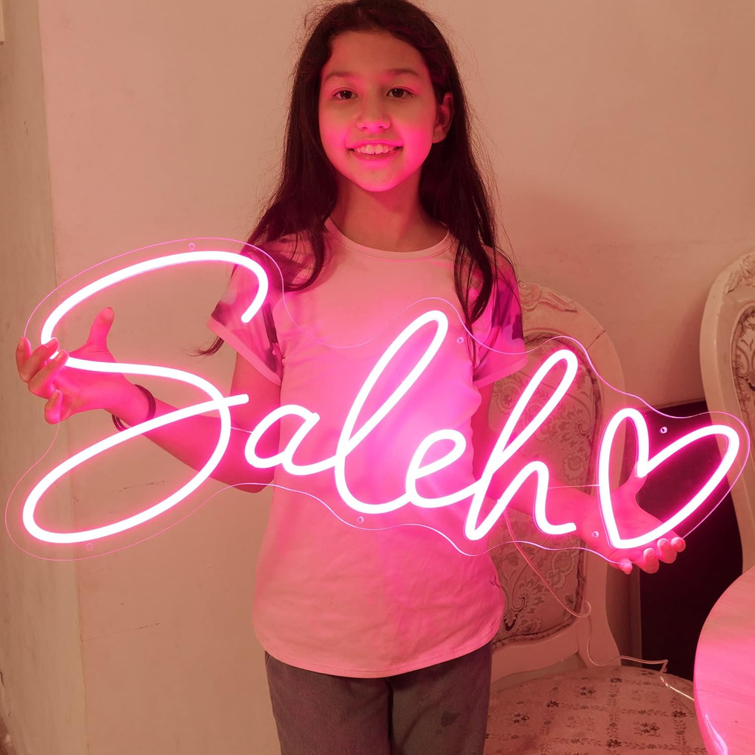 A young girl holds a bright pink neon sign displaying the name \“Saleh\“ with a heart design.