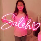 A young girl holds a bright pink neon sign displaying the name \“Saleh\“ with a heart design.