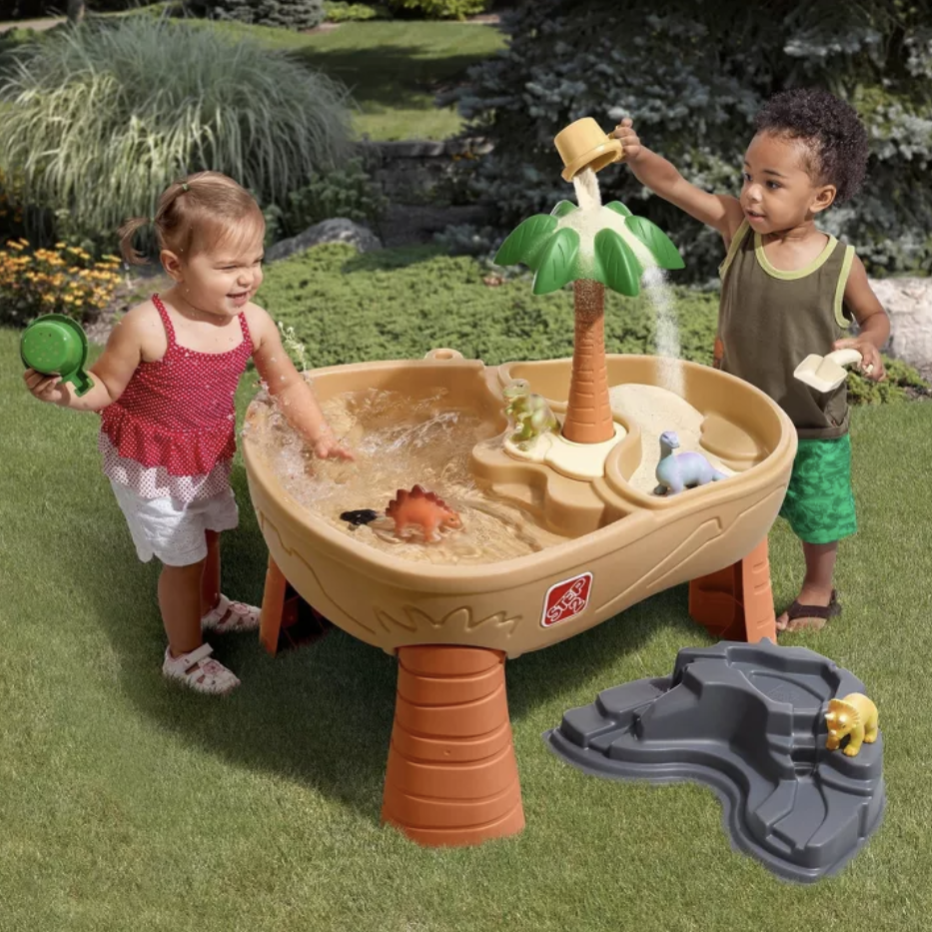 Children play with a sand and water activity table featuring a palm tree, dinosaur figurines, and a pouring cup. The setup includes a separate rocky water area, providing interactive play experiences outdoors on grass.