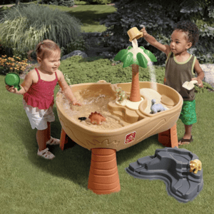 Children play with a sand and water activity table featuring a palm tree, dinosaur figurines, and a pouring cup. The setup includes a separate rocky water area, providing interactive play experiences outdoors on grass.