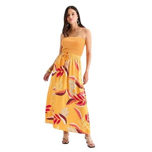 A woman is wearing a strapless yellow maxi dress with a colorful floral pattern and accessorized with hoop earrings and brown sandals.