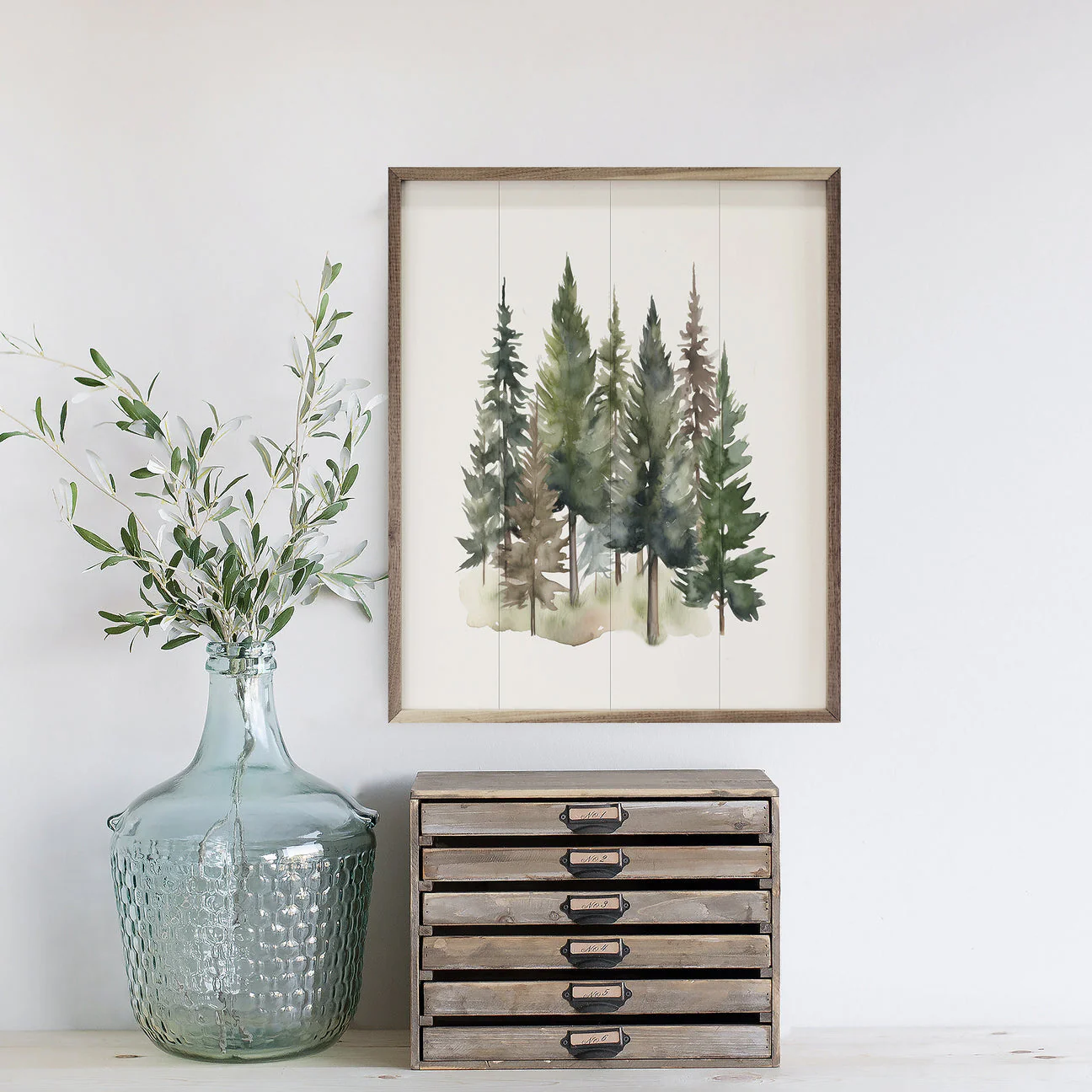 A watercolor painting of evergreen trees in a wooden frame is displayed on a wall. Below is a large textured glass jar containing leafy branches, next to a small wooden drawer unit.