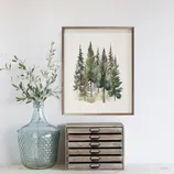 A watercolor painting of evergreen trees in a wooden frame is displayed on a wall. Below is a large textured glass jar containing leafy branches, next to a small wooden drawer unit.