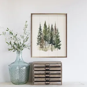 A watercolor painting of evergreen trees in a wooden frame is displayed on a wall. Below is a large textured glass jar containing leafy branches, next to a small wooden drawer unit.
