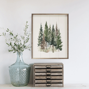 A watercolor painting of evergreen trees in a wooden frame is displayed on a wall. Below is a large textured glass jar containing leafy branches, next to a small wooden drawer unit.