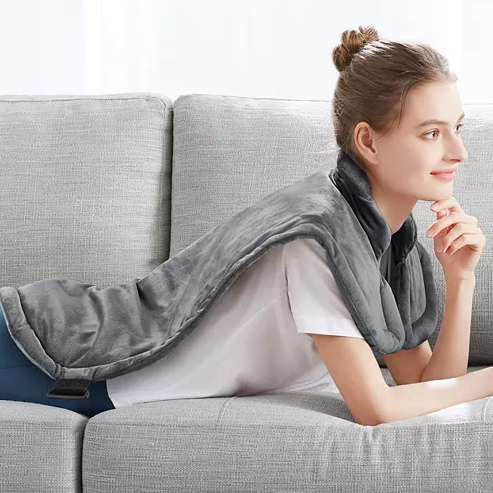 A person is reclining on a couch with a plush, gray wrap draped around their neck and extending down their back, designed for providing heat.
