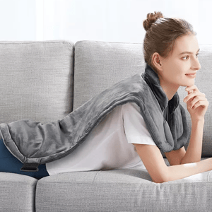 A person is reclining on a couch with a plush, gray wrap draped around their neck and extending down their back, designed for providing heat.