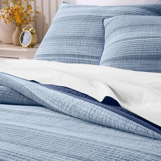 A neatly made bed features a light blue quilted cover and matching pillows with a white sheet partially exposed underneath. A gold-colored alarm clock and yellow flowers are on the nightstand.