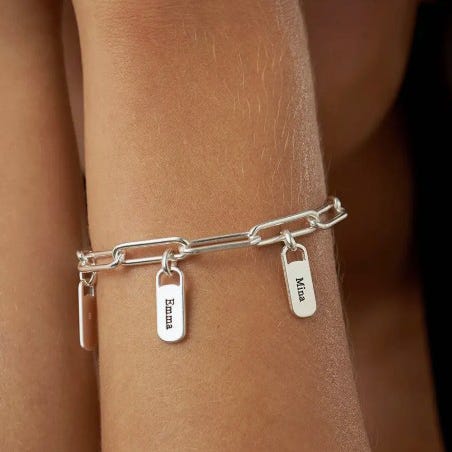A silver bracelet with rectangular name charms hangs on an arm, featuring engraved names like Emma and Mila.