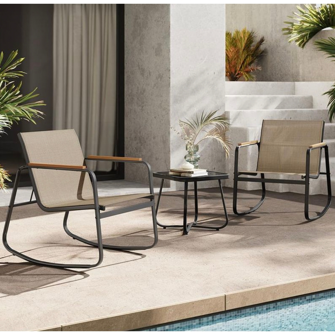 3-piece patio set includes two beige mesh rocking chairs with black metal frames and wooden armrests, paired with a small black round side table. Decorated with plants, the setup is situated next to a pool on a stone patio.