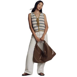 A woman wears a striped sleeveless top and loose white pants, holding a large brown bag. She pairs the outfit with dark sandals.