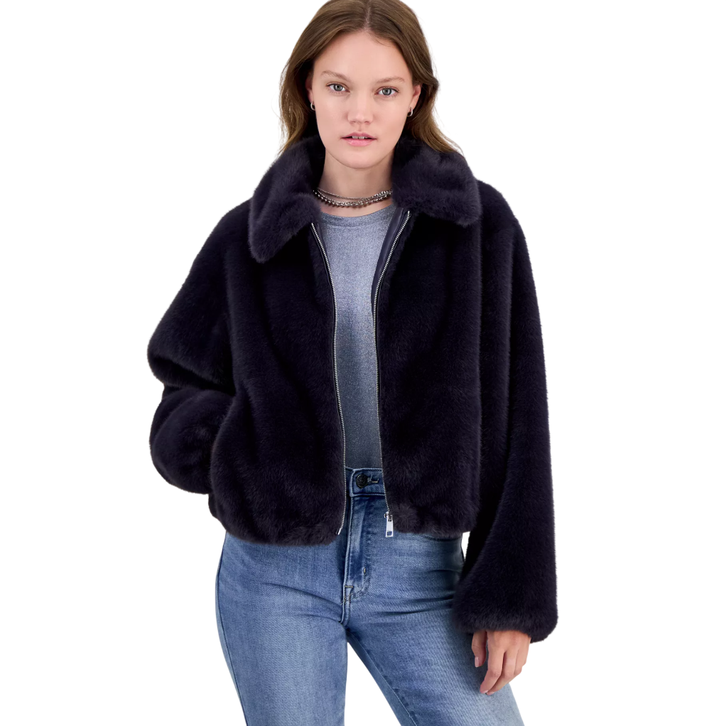 Woman wearing a dark faux fur jacket, gray shirt, and blue jeans, accessorized with a silver necklace.