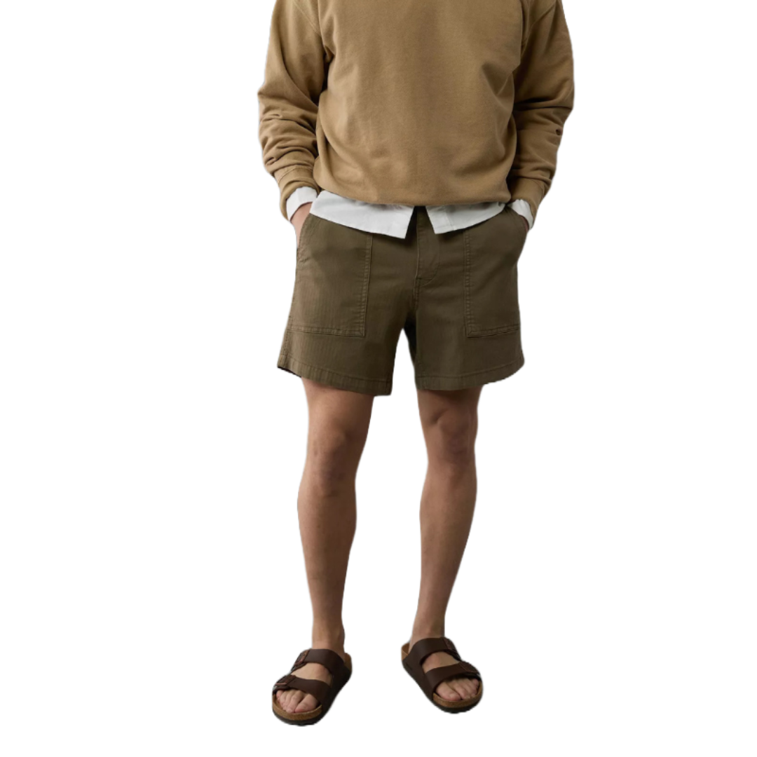 The Flex Utility Short is an olive green, knee-length short featuring large front patch pockets. It's paired with brown sandals, a beige sweatshirt, and a white shirt underneath.