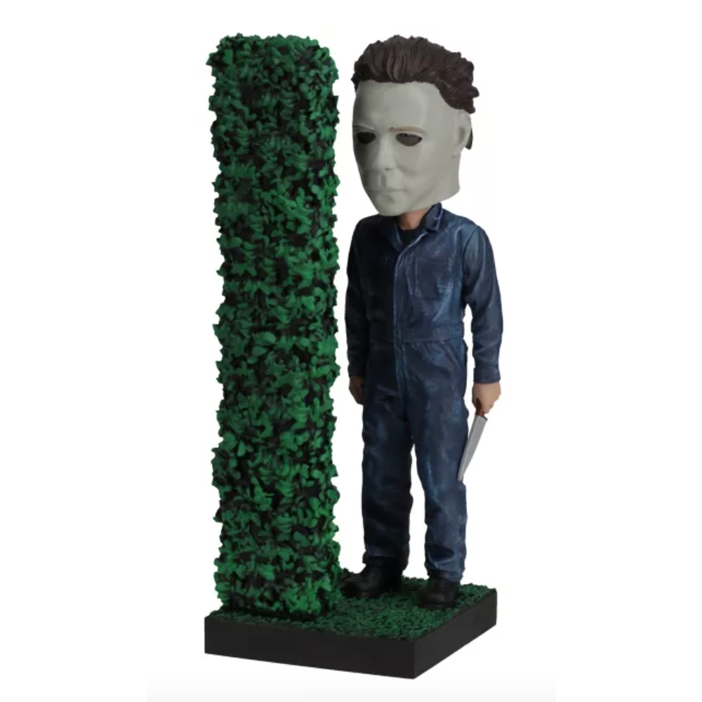A Michael Myers bobblehead features the character in a blue jumpsuit and mask, standing next to a green bush on a black base.
