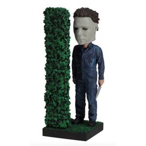 A Michael Myers bobblehead features the character in a blue jumpsuit and mask, standing next to a green bush on a black base.