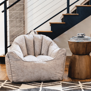 A cozy gray bean bag chair sits beside a wooden side table with books stacked on top. The scene is set in a modern interior with a geometric patterned rug and a staircase in the background.