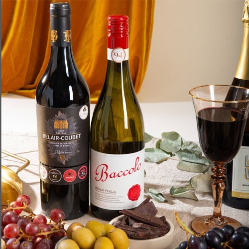 Two wine bottles, Belair-Coubet and Baccorlo, are displayed on a table with grapes, olives, chocolate, a glass of wine, and a bottle of sparkling wine in the background.