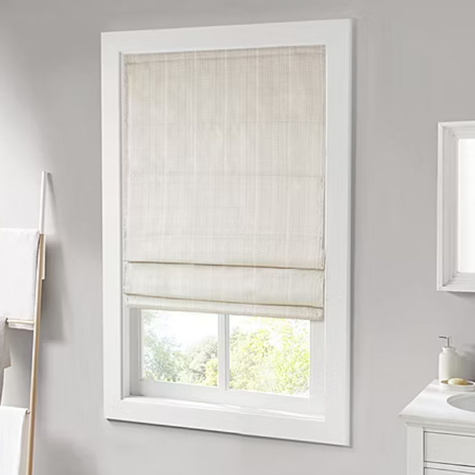 Light-colored Roman shade covering a rectangular window, surrounded by a white frame in a gray-walled room. A partial view of a towel rack and bathroom sink is visible nearby.