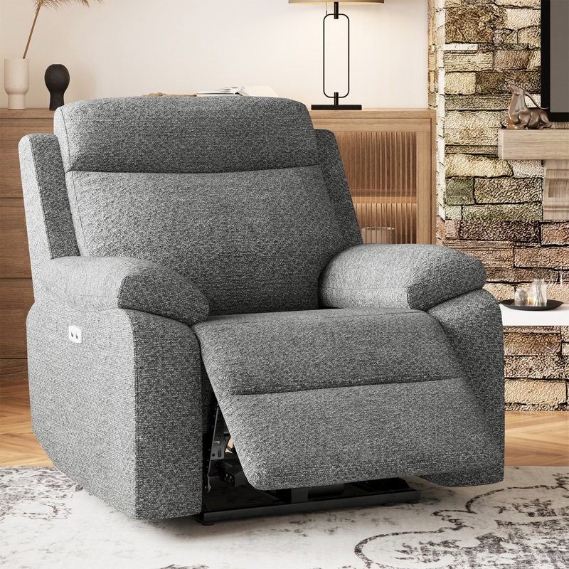 A gray upholstered reclining armchair with cushioned armrests and an extended footrest, positioned in a cozy living room setting with a wooden cabinet and a brick fireplace in the background.