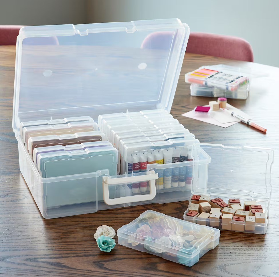 A clear plastic storage case with multiple smaller cases inside, each holding an assortment of colorful craft supplies, like stamps and markers, on a wooden table.