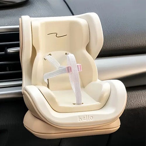 A miniature car seat-style phone holder is attached to a vehicle's air vent, featuring a beige and cream design with a mock seatbelt.