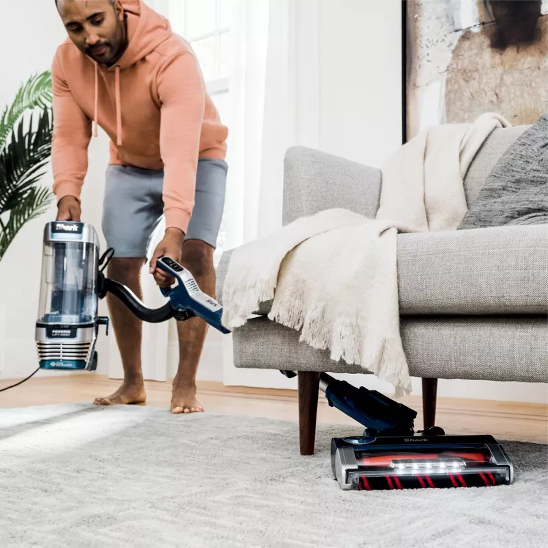 A man is vacuuming a carpet using an upright Shark vacuum cleaner.