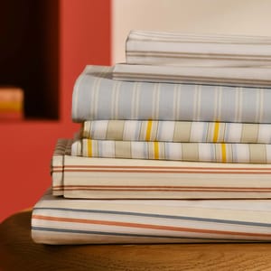 A stack of folded bed sheets in various striped patterns and colors, including blue, yellow, green, and orange, neatly arranged on a wooden surface.