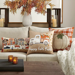 Autumn-themed decorative pillows with a pumpkin design, a truck carrying pumpkins, and a town scene, complemented by a knit throw and candle holders.
