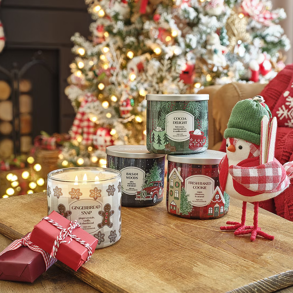 Scented candles with seasonal aromas and a festive bird decoration on a wooden table, with a Christmas tree in the background.