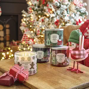 Scented candles with seasonal aromas and a festive bird decoration on a wooden table, with a Christmas tree in the background.