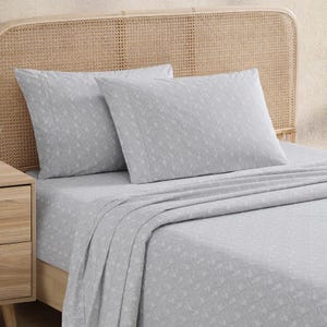 Tommy Bahama Percale Queen Sheet Sets feature a subtle, elegant leaf pattern in light gray, made from soft percale cotton. The set includes pillowcases, a flat sheet, and a fitted sheet, displayed on a wooden bed with a woven headboard.