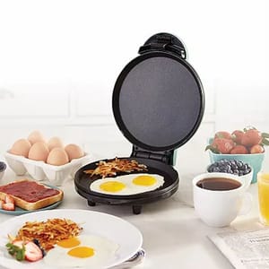 An open, round, black electric griddle cooking two eggs, with a breakfast setup including a plate of eggs, toast, fruit, and coffee.