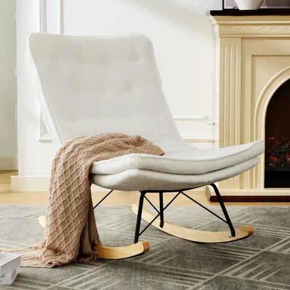 A white, armless rocking chair features a cushioned, tufted seat and a sleek black metal frame with wooden rockers. A beige, textured throw is draped over the chair, and it's positioned on a patterned gray rug near a fireplace.
