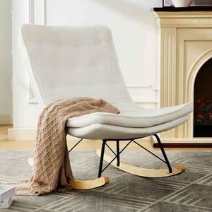 A white, armless rocking chair features a cushioned, tufted seat and a sleek black metal frame with wooden rockers. A beige, textured throw is draped over the chair, and it's positioned on a patterned gray rug near a fireplace.