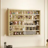 A wooden wall-mounted display case holds various collectible figurines and toys, including animal figures, action characters, and mini bikes, with a glass front. Below, a light-colored cabinet features a candles, a bowl, and a space-themed figure.
