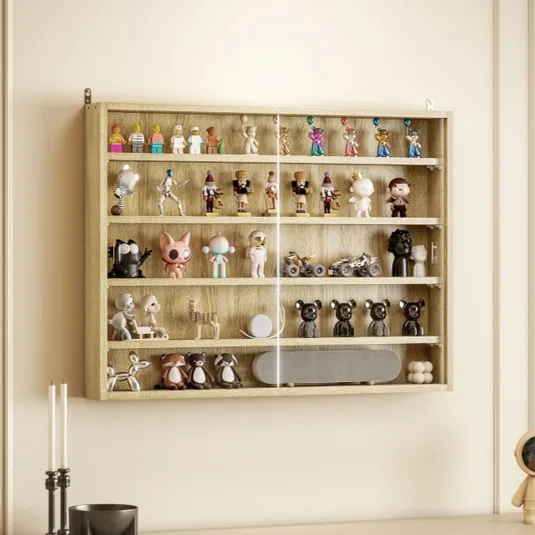 A wooden wall-mounted display case holds various collectible figurines and toys, including animal figures, action characters, and mini bikes, with a glass front. Below, a light-colored cabinet features a candles, a bowl, and a space-themed figure.