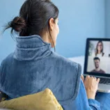 A person uses a heating pad draped over their shoulders while attending a video call on a laptop.