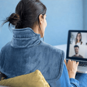 A person uses a heating pad draped over their shoulders while attending a video call on a laptop.