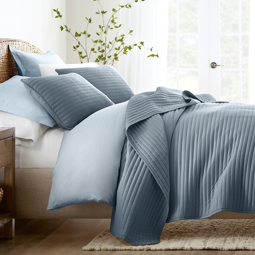 A bed with light and dark blue pleated bedding, including a comforter, pillows, and sheets, set in a bright room with a plant in the background.