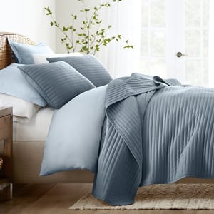 A bed with light and dark blue pleated bedding, including a comforter, pillows, and sheets, set in a bright room with a plant in the background.