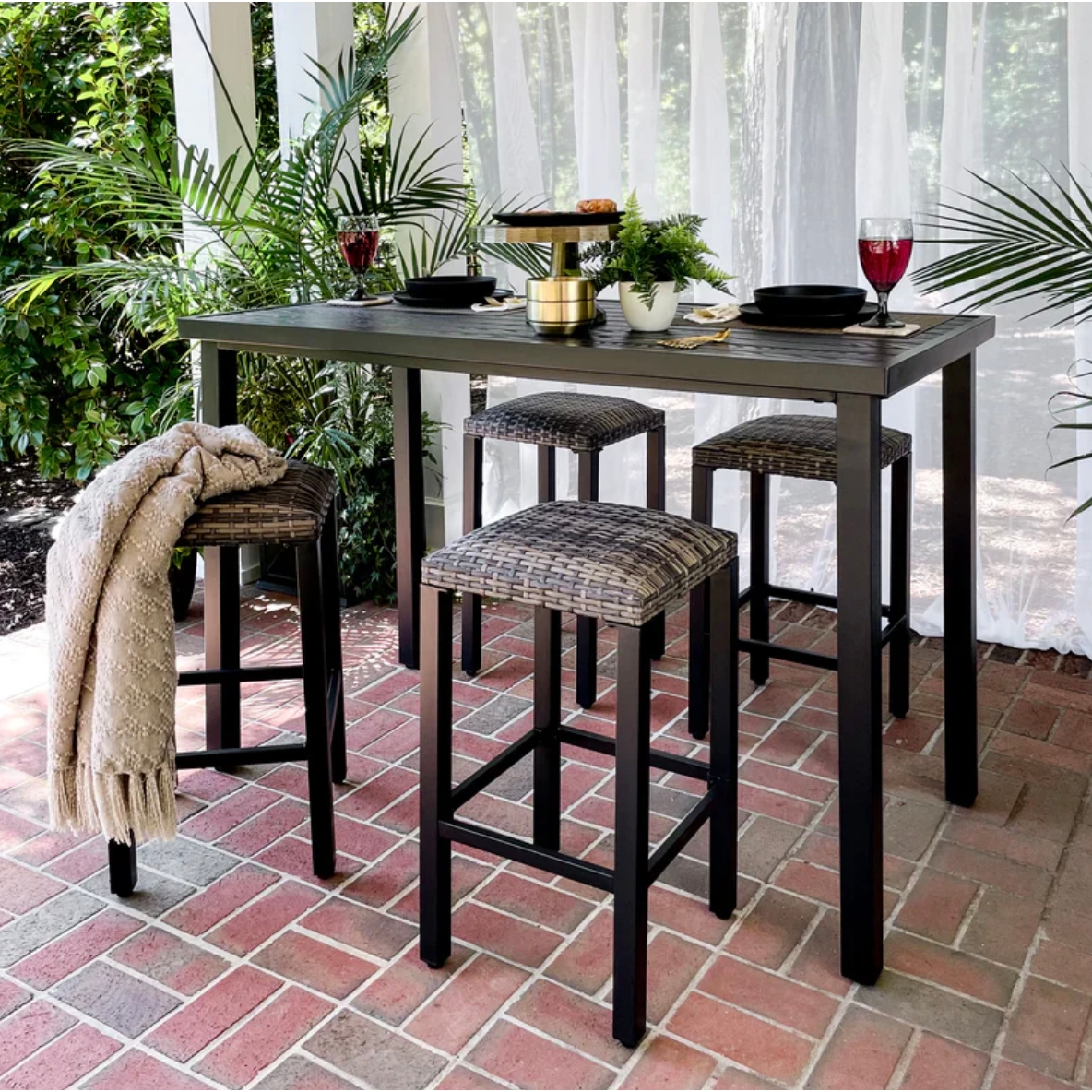 Outdoor bar-height table set with four woven stools on a brick patio; includes black plates, glasses, a tiered serving tray, and potted plants under a sheer curtain canopy.