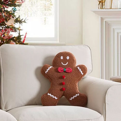 A gingerbread man-shaped pillow with a smiling face and white and red decorative details sits on a chair.
