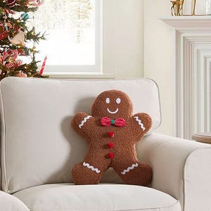 A gingerbread man-shaped pillow with a smiling face and white and red decorative details sits on a chair.