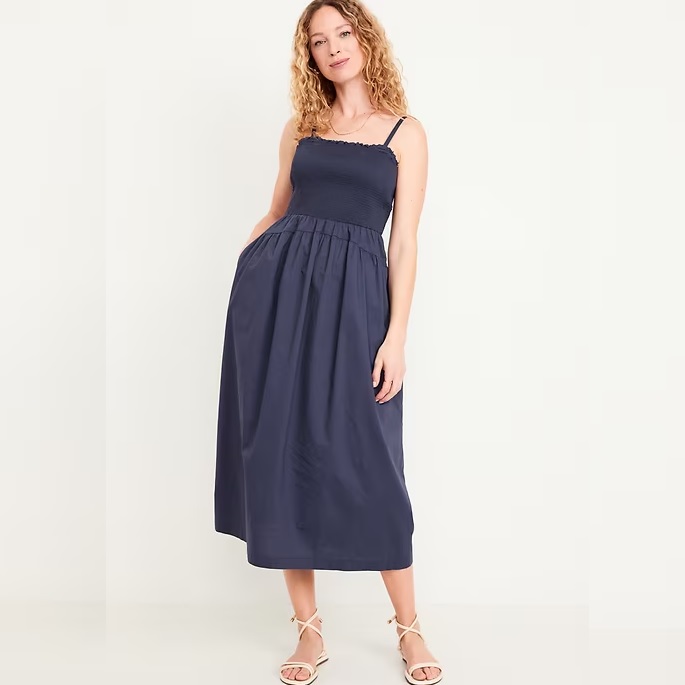 A woman models a navy blue midi dress with a smocked bodice and spaghetti straps, paired with light beige strappy sandals.