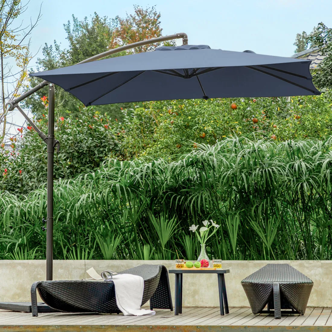 A poolside scene with a large patio umbrella, two wicker lounge chairs, and a small table holding drinks and fruit, set against lush greenery.