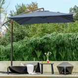 A poolside scene with a large patio umbrella, two wicker lounge chairs, and a small table holding drinks and fruit, set against lush greenery.