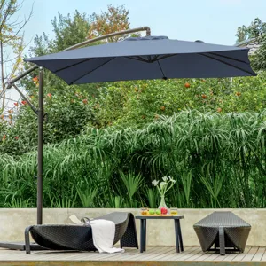 A poolside scene with a large patio umbrella, two wicker lounge chairs, and a small table holding drinks and fruit, set against lush greenery.