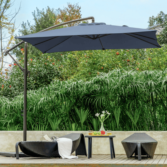 A poolside scene with a large patio umbrella, two wicker lounge chairs, and a small table holding drinks and fruit, set against lush greenery.