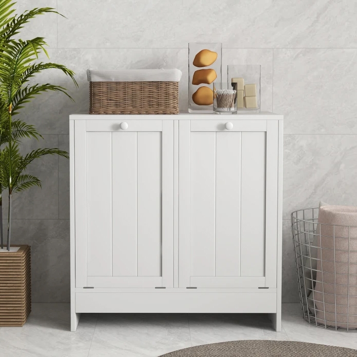 A white, double tilt-out laundry hamper with vertical panel doors and round knobs, topped with a wicker basket and decorative items. It stands beside a wire basket and a potted plant on a tiled floor.