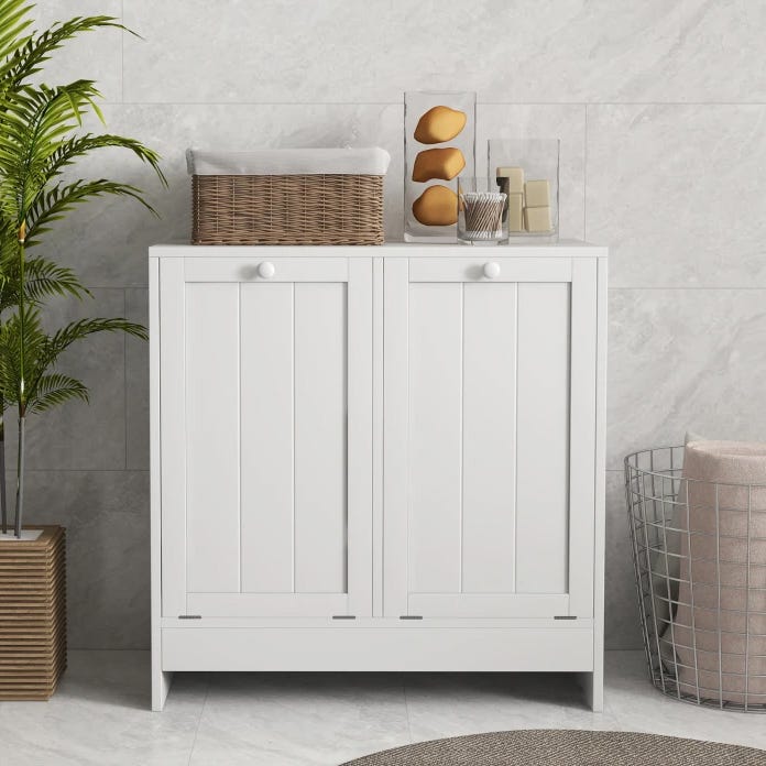 A white, double tilt-out laundry hamper with vertical panel doors and round knobs, topped with a wicker basket and decorative items. It stands beside a wire basket and a potted plant on a tiled floor.